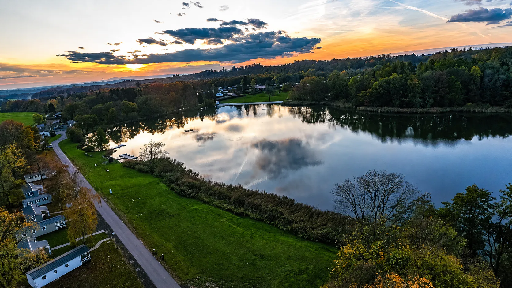 Luftaufnahme des Gederner Sees bei Sonnenuntergang mit Mobilheimen am Ufer und farbigem Himmel